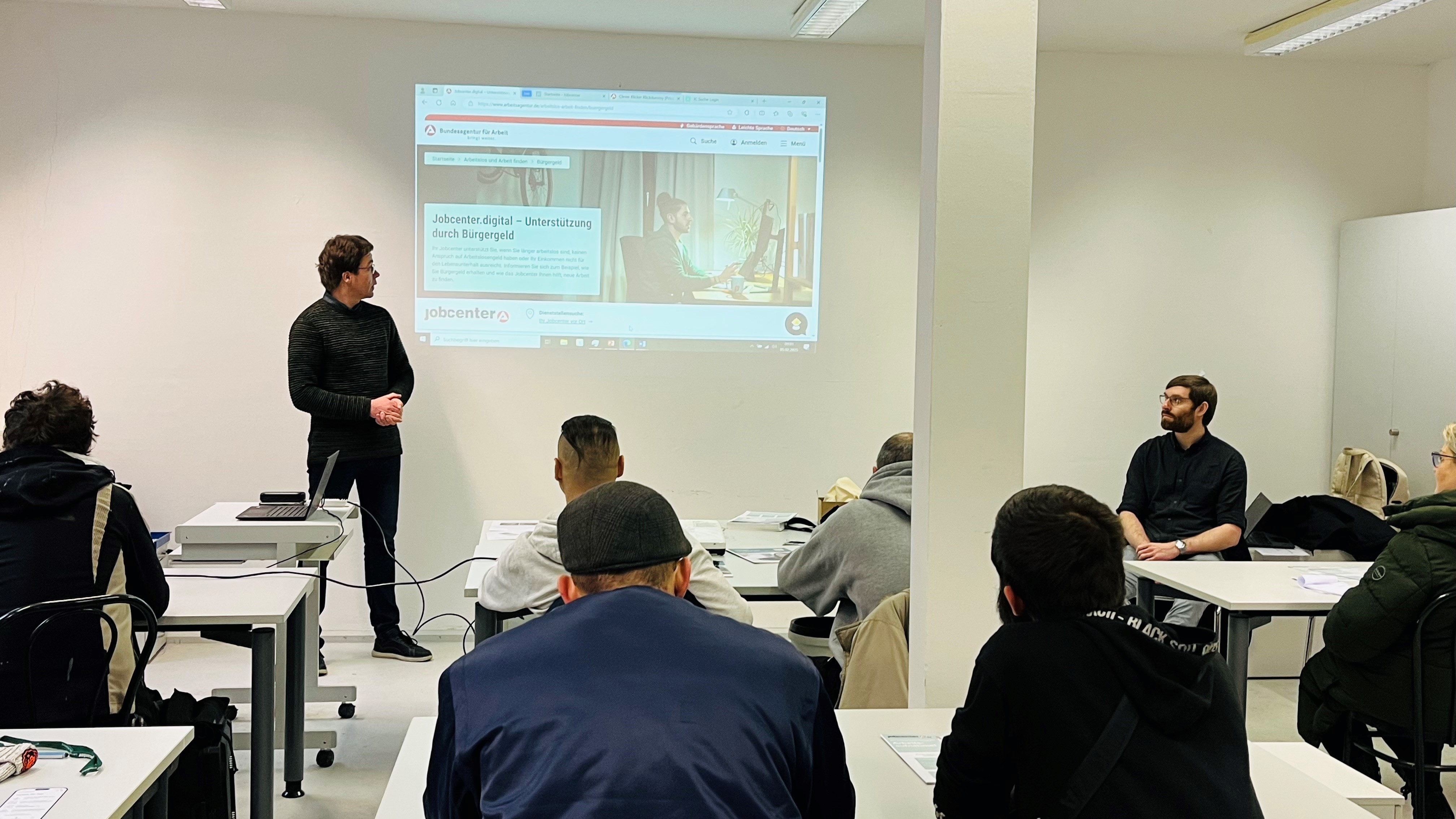 Zu sehen sind zwei Jobcenter Mitarbeiter, die vor einer Gruppe Menschen steht. Im Hintergrund wird die online Plattform von www.jobcenter.digital mittels Beamer auf der Wand gezeigt. Zu sehen sind zwei Jobcenter Mitarbeiter, die vor einer Gruppe Menschen steht. Im Hintergrund wird die online Plattform von www.jobcenter.digital mittels Beamer auf der Wand gezeigt.